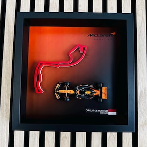 Peut inclure: Œuvre d'art encadrée représentant une voiture McLaren de Formule 1 et un tracé rouge du Circuit de Monaco. L'œuvre comprend le logo McLaren et le texte "CIRCUIT DE MONACO". La voiture est noire et orange.