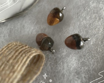 May include: Three brown and black acorn-shaped charms with silver loops. The charms are on a light gray surface.