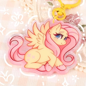 May include: A yellow and pink acrylic keyring featuring a cartoon pony with pink hair and wings. The keyring has a yellow heart-shaped clasp. The text "@MOONLIGHTTDREAMS" is at the bottom of the keyring.