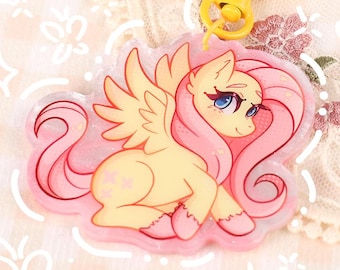 Puede incluir: Un llavero acrílico amarillo y rosa con un pony de dibujos animados con pelo y alas rosas. El llavero tiene un cierre amarillo en forma de corazón. El texto "@MOONLIGHTTDREAMS" está en la parte inferior del llavero.