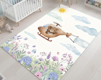 May include: A whimsical children's rug featuring a brown teddy bear piloting a helicopter, with a banner that says "Hello". The rug has a floral border with lavender, blue, and pink flowers, and a sky with clouds and a sun.