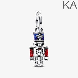 May include: A silver charm in the shape of a toy soldier nutcracker. The charm has a blue and red colour scheme with a star on the hat. The arms are red with glitter. The charm is a festive holiday decoration.