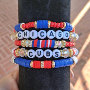 May include: Stack of beaded bracelets in red, white, blue, and gold. The bracelets spell out "CHICAGO CUBS" with white letter beads. The bracelets are stacked on a black cylindrical surface.