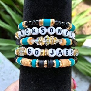 May include: A stack of four beaded bracelets featuring black, teal, and tan clay discs, gold-tone spacers, and clear crystal accents. Two bracelets include white letter beads spelling out JACKSONVILLE and GO JAGS in black capital letters.