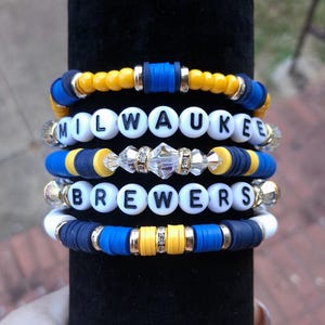 May include: Stack of five beaded bracelets. The bracelets feature white letter beads spelling "MILWAUKEE BREWERS", interspersed with blue, yellow, and gold beads. Some bracelets have clear crystal accents. The bracelets are displayed on a black cylindrical stand.