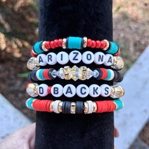 May include: Stack of beaded bracelets with the words "ARIZONA" and "O BACKS" spelled out in white letter beads. The bracelets feature red, turquoise, black, and gold accents. The bracelets are stacked on a black velvet surface.
