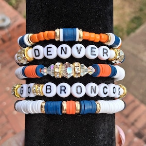 May include: Five stacked bracelets with white, orange, and navy blue beads. Two bracelets spell out "DENVER" and "GO BRONCOS" with white letter beads. Gold and crystal accents are also present.