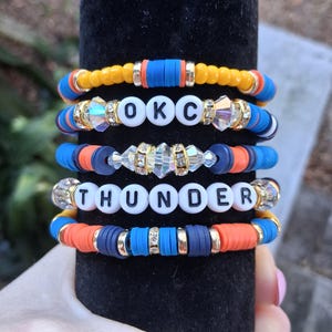May include: Stack of five beaded bracelets in yellow, blue, orange, and clear beads. Two bracelets spell out "OKC THUNDER" in white letter beads. The bracelets are stacked on a black velvet surface.