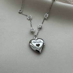May include: A silver heart-shaped locket necklace with a pearl chain. The locket has the initials "WGS" engraved on it and a small heart inside.