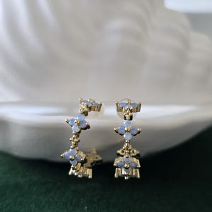 May include: A pair of gold-toned earrings featuring a floral design with light blue gemstones. The earrings have a post-back closure and a delicate, elegant appearance. The earrings are displayed against a soft, blurred background.