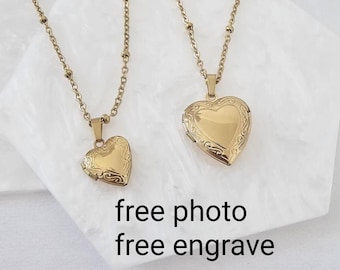May include: Two gold-toned heart-shaped lockets on delicate beaded chains. Each locket features an ornate engraved border and a smooth center. The text "free photo free engrave" is visible below the jewelry pieces on the white surface.