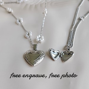May include: Two silver heart-shaped locket necklaces. One necklace has a delicate chain with small white flower charms. The other necklace has a thicker chain. The text "free engrave, free photo" is written below the necklaces.