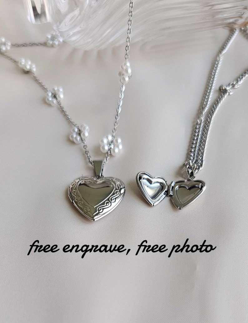 May include: Two silver heart-shaped locket necklaces. One necklace has a delicate chain with small white flower charms. The other necklace has a thicker chain. The text "free engrave, free photo" is written below the necklaces.