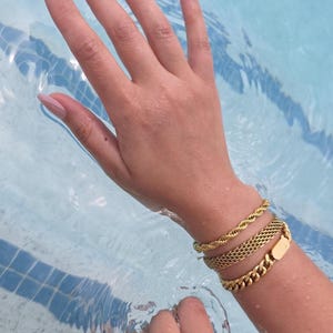 May include: Gold bracelets in various styles, including a rope chain, a mesh design, and a curb chain, are displayed on a wrist in a pool. The water is a light blue color, and the hand has long, manicured nails.