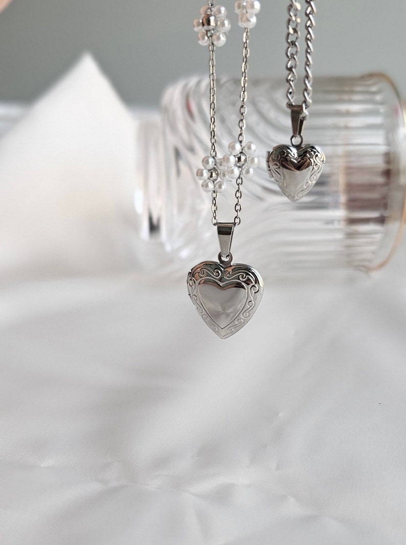 May include: Two silver heart-shaped locket necklaces. One necklace has a delicate chain with small pearl-like beads. The other necklace has a thicker chain.
