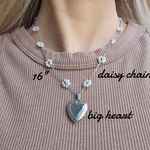 May include: A silver necklace with a large heart pendant and a daisy chain design. The necklace is 16 inches long and features a delicate silver chain with small white flower accents. The heart pendant has a detailed design and is a statement piece.