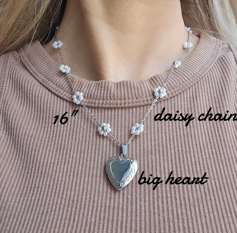 May include: A silver necklace with a large heart pendant and a daisy chain design. The necklace is 16 inches long and features a delicate silver chain with small white flower accents. The heart pendant has a detailed design and is a statement piece.
