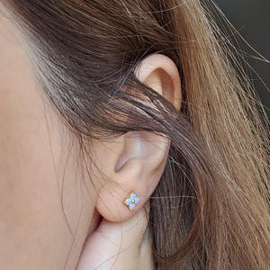 May include: A small gold stud earring shaped like a four-petaled flower with light blue, iridescent stone inlays. The earring is centred on the earlobe, featuring a metallic gold frame that outlines each petal and a small gold bead at the centre.