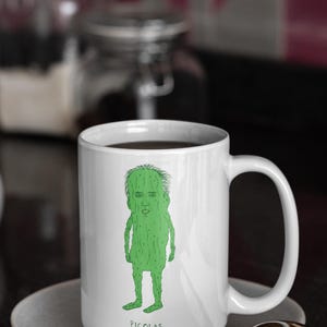 Könnte beinhalten: Weiße Keramiktasse mit einer grünen Cartoon-Illustration von Nicolas Cage als Gurke. Die Tasse ist mit einer dunklen Flüssigkeit gefüllt und steht auf einer grauen Untertasse. Der Text "PICOLAS CAGE" ist darunter gedruckt.