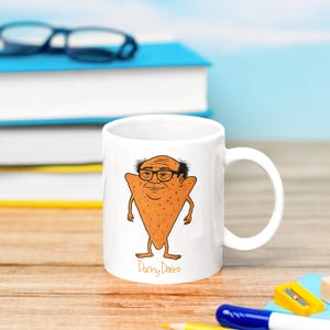 May include: White ceramic mug featuring a cartoon illustration of a person with glasses and a triangle-shaped body. The mug has the text "Danny DeVito" at the bottom. The background includes books, pencils, and a pencil sharpener.