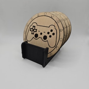 May include: Set of six wooden coasters featuring a laser-etched design of a video game controller. The coasters are stacked and displayed in a black stand.