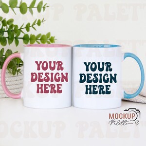 Op de afbeelding: Twee witte keramische mokken met gekleurde randen. De mok links heeft een roze rand en de tekst "YOUR DESIGN HERE" in donkerrood. De mok rechts heeft een blauwe rand en de tekst "YOUR DESIGN HERE" in donkerblauw.