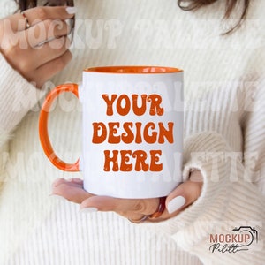May include: White ceramic mug with an orange interior, handle, and the text "YOUR DESIGN HERE" in orange. The mug is held by a person wearing a white sweater. The mug is a standard size for coffee or tea.