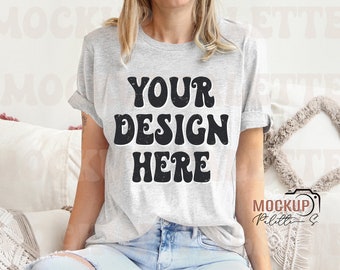May include: A gray t-shirt with the text "YOUR DESIGN HERE" in black retro-style lettering. The shirt is being modeled by a person wearing blue jeans.