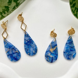 Könnte beinhalten: Ein Paar blaue und goldene Tropfenohrringe mit Marmoreffekt. Die Ohrringe haben eine goldene Tropfenform mit einer goldenen Scheibe oben.