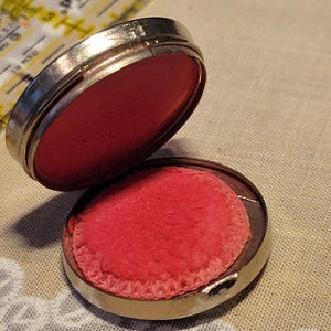 May include: Open antique silver compact with a bright red powder puff. The compact is round and has a hinged lid. The powder puff is a circular shape with a decorative edge. The compact is open, revealing the red powder.