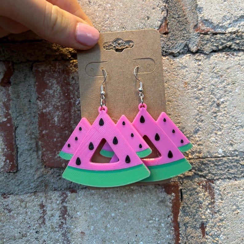 May include: A pair of pink watermelon slice earrings with black seeds and a green rind. The earrings are hanging from silver hooks.
