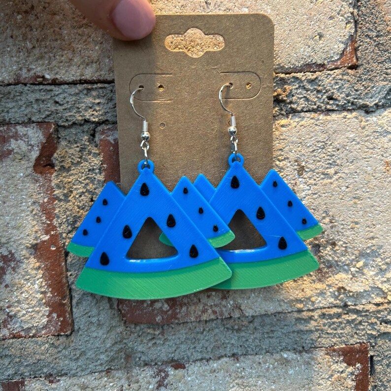 May include: A pair of watermelon slice earrings. The earrings are made of blue, green, and black acrylic. The earrings are hanging from silver hooks.