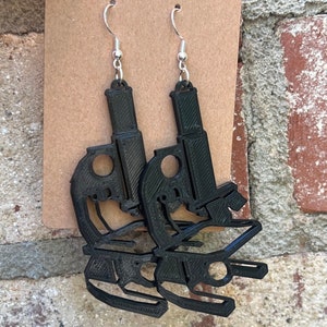 May include: A pair of black microscope earrings with silver-colored ear wires. The earrings are 3D-printed and have a detailed design, showcasing the shape of a microscope. The earrings are hanging against a brown card.