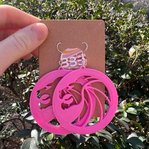 May include: Pink plastic earrings shaped like a lion's head with a wave design. The earrings are hanging from silver hooks.