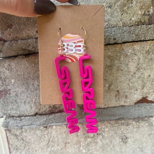 May include: A pair of pink dangle earrings with the word "DREAM" spelled out in a 3D font. The earrings are on a brown card with a sticker that says "333 Prints".