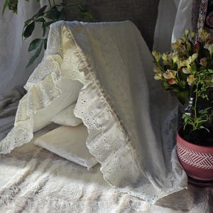 Puede incluir: Tela crema con volantes y calados, drapeada sobre dos almohadas blancas. La tela presenta un delicado diseño calado. Un jarrón con flores y una planta en maceta son visibles en el fondo, sobre una superficie de piel sintética.