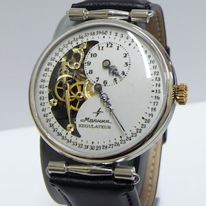 May include: A silver-toned wristwatch with a black leather strap and a white dial featuring a regulator display. The dial shows a minute track from 1 to 60, a sub-dial for hours, and an exposed gold-colored mechanical movement. Text reads Molnija.