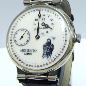 May include: A silver-toned wristwatch with a white dial featuring a black leather strap. The dial displays the text MEMENTO MORI, a grim reaper illustration, a sub-dial, and a numbered outer ring from 1 to 60. The Omega logo is printed in black.
