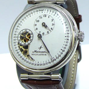 May include: A silver-toned wristwatch with a white dial, featuring a central hour sub-dial, a large minute hand, and a skeletonized seconds sub-dial. The dial displays the text "Молния" and "REGULATEUR" with a minute track numbered 1 to 60.
