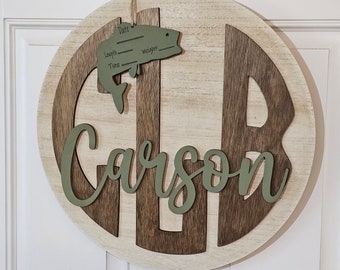 May include: A round wooden sign with a brown and white wood grain pattern. The sign has a large green "C" in the center, with the name "Carson" written in green letters below. A small green fish cutout with a blank space for writing "Date", "Length", "Weight", and "Time" is hanging from the top of the sign.