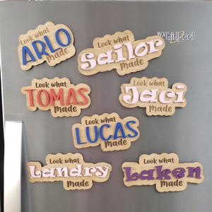 May include: Wooden name magnets with the phrases "Look what Made" and various names in different colors: Arlo, Sailor, Tomas, Jaci, Lucas, Landry, and Laken. The magnets are attached to a gray refrigerator.