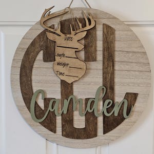 Puede incluir: Un letrero redondo de madera con el nombre "Camden" en escritura verde. El letrero presenta un recorte de cabeza de ciervo con espacio para la fecha, la longitud, el peso y la hora. El fondo es de vetas de madera clara.