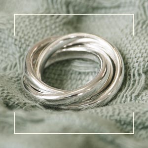 May include: A silver-colored ring with five interlocking bands. The ring is made of a shiny metal and is displayed on a textured, light green surface. The bands are smooth and rounded, creating a continuous, intertwined design.