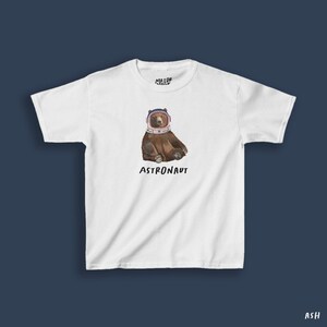 May include: White t-shirt featuring a brown bear wearing an astronaut helmet. The word "ASTRONAUT" is printed below the bear. The shirt has a crew neck and short sleeves. The brand name "MAJOR ASHLEY" is printed at the top.