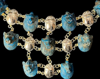 Puede incluir: Un collar de cadena dorada con colgantes azules y plateados. Los colgantes presentan la imagen de un escarabajo y una cabeza de gato.