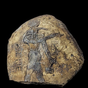Puede incluir: Una talla de piedra de un dios egipcio con cabeza de halcón, sosteniendo un bastón y un ankh. La talla está sobre una piedra áspera e irregular. El nombre del dios, "Horus", está inscrito en jeroglíficos en el lado izquierdo de la talla.
