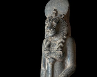 May include: A dark gray stone statue of an Egyptian deity with a lion head and a crown. The statue is carved with intricate details and has a weathered appearance.