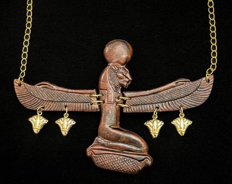 Puede incluir: Un collar de cadena dorada con un colgante de madera marrón que representa a una diosa egipcia con las alas extendidas. El colgante tiene cinco pequeños amuletos de oro en forma de flor.
