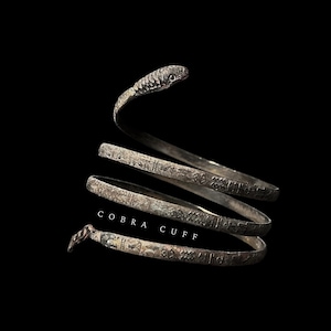 May include: A silver cobra cuff bracelet with intricate details. The bracelet is coiled into a spiral shape with the head of the cobra at the top. The text "COBRA CUFF" is printed on the bracelet.