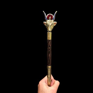 Puede incluir: Un bastón dorado con un remate de cabeza de toro. La cabeza de toro tiene una gema roja en el centro de la frente y detalles de esmalte azul alrededor de los ojos. El bastón está hecho de madera oscura.